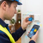 Zbliżenie na elektryka w niebieskim mundurze, kasku i żółtej kamizelce odblaskowej, który instaluje mały niebieski przekaźnik smart home do puszki elektrycznej w ścianie. Jednocześnie trzyma w lewej ręce smartfon z uruchomioną polskojęzyczną aplikacją "Dom Mój" (Mój Dom). Od zainstalowanego przekaźnika i smartfona rozchodzą się stylizowane fale sygnału bezprzewodowego. Na ekranie telefonu widoczne są ikony: żarówka z literą "A" (Światło), słońce (Sceny), dom i profil użytkownika. W tle widać fragment salonu z drewnianą szafką i rośliną doniczkową.